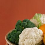 Close-up of fresh broccoli, cauliflower, lemon, and orange on a wooden plate with an earthy background.